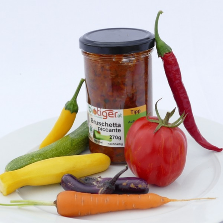 Ein weißer Teller enthält ein Glas Bruschetta Piccante, eine rote Tomate, eine Karotte, Auberginen, gelbe Zucchini und rote und gelbe Paprikaschoten.