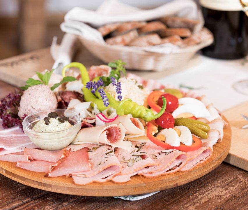 Ein Holztablett mit einer Auswahl an Fleisch, Käse und Gemüse, begleitet von einer Schüssel Dip und einem Korb mit Brot.