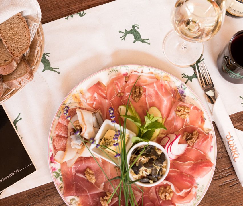 Ein Holztisch ist mit einem Teller mit Fleisch, Nüssen, Gemüse und Kräutern, begleitet von einem Korb mit Brot, einem Weinkelch und einer Flasche gedeckt.