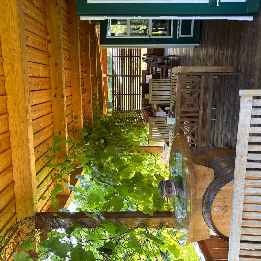 Eine hölzerne Veranda mit Blick auf eine üppige grüne Pflanze, einen Fasstisch und einen Holzstuhl. Es gibt auch Holzbretter an der Wand und ein Fenster.