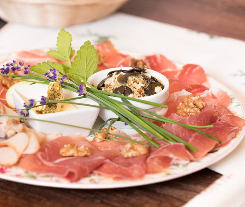Ein Teller mit dünn geschnittenem Prosciutto, überzogen mit einer Schüssel mit Nüssen, Käse und einem Zweig Minze. Der Teller hat auch einige grüne Blätter und lila Blumen.