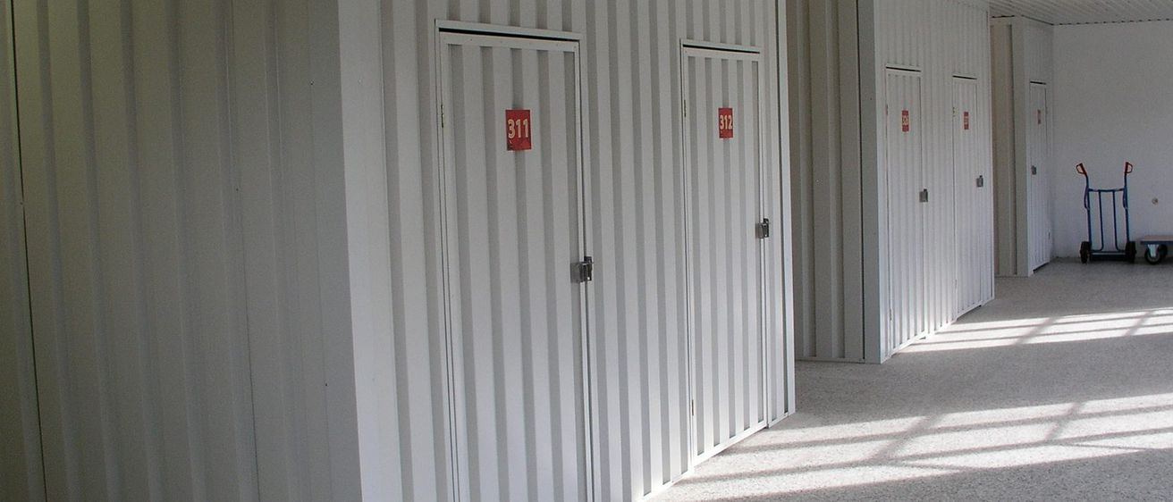 Ein leerer Lagerraum mit weißen Wänden und Metallverkleidung, mit '311' auf der Tür beschriftet. Der Boden ist grau und sauber, mit einem roten Trolley draußen geparkt.