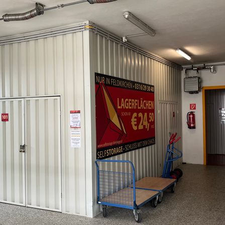 Ein Lagerraum mit einem roten Schild, das Lagerflächen für 24,50 € pro Monat anbietet. Es gibt zwei Wagen und einen Feuerlöscher an der Wand. Die Einheitsnummer 101 befindet sich auf der Tür.