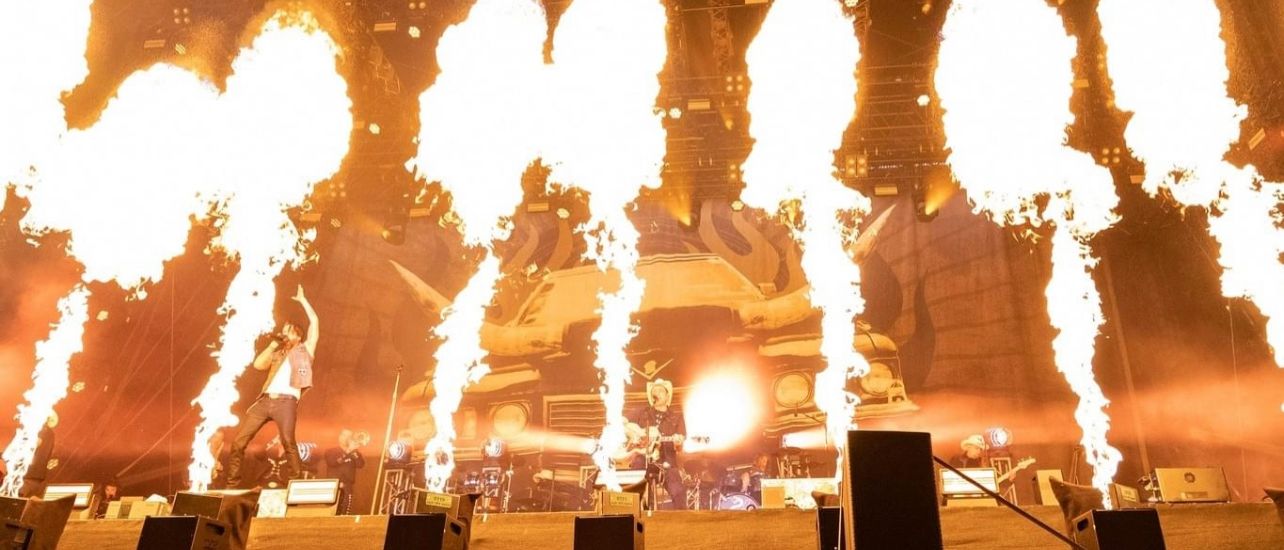 Ein Live-Konzert auf der Bühne mit Feuereffekten, einem Schlagzeuger und einem Gitarristen. Die Bühne ist mit schwarzem Stoff bedeckt und das Publikum schaut zu.