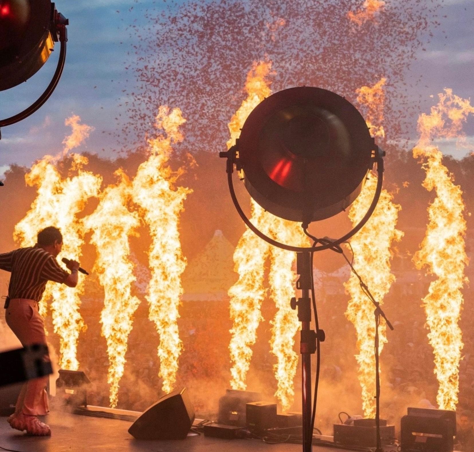 Ein Performer auf der Bühne mit Mikrofon, umgeben von hohen Flammen. Ein großer Scheinwerfer ist vor ihm positioniert und beleuchtet die Szene.