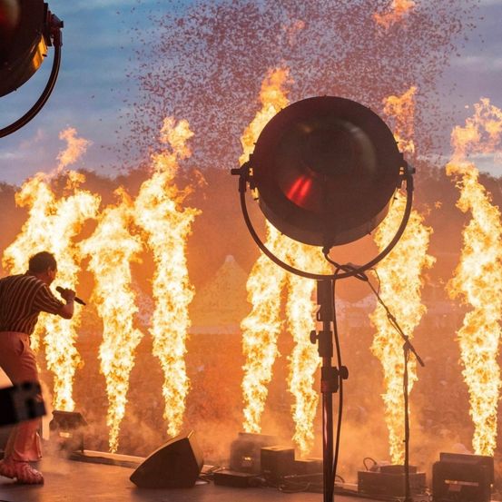 Ein Künstler steht mit einem Mikrofon auf der Bühne, mit großen, flammenden Fackeln im Hintergrund. Scheinwerfer und Lautsprecher sind um die Bühne herum positioniert, mit einer Menge im Hintergrund.