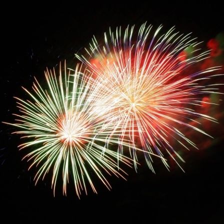 Zwei Feuerwerke im Nachthimmel. Eines sendet grüne und weiße Funken aus, das andere rote und gelbe. Die Nacht ist dunkel, das einzige Licht kommt von den Feuerwerken.