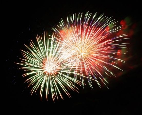 Zwei Feuerwerke im Nachthimmel. Eines sendet grüne und weiße Funken aus, das andere rote und gelbe. Die Nacht ist dunkel, das einzige Licht kommt von den Feuerwerken.