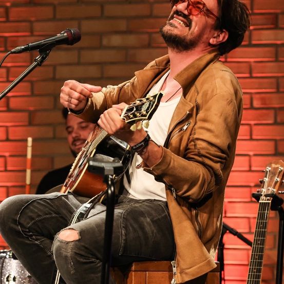 Ein Mann in einer braunen Jacke und zerrissenen Jeans spielt Gitarre auf der Bühne. Er sitzt mit einem Mikrofon vor einer Ziegelwand.