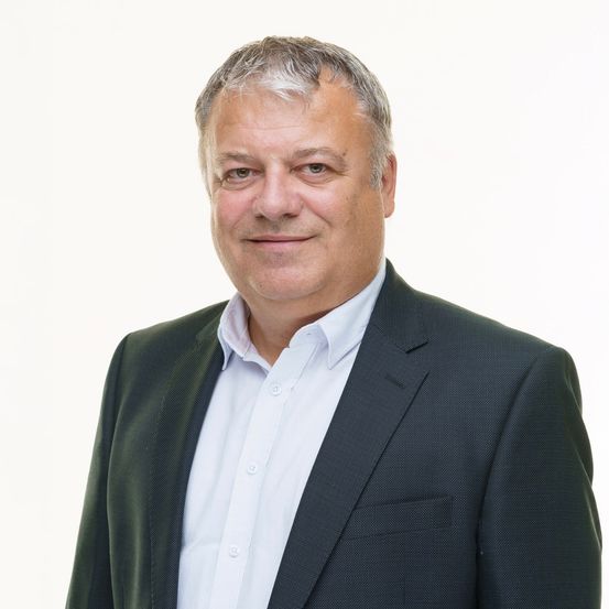 Ein Mann in einem Anzug mit grauem Haar lächelt direkt in die Kamera. Er trägt ein weißes Hemd und einen dunklen Blazer. Der Hintergrund ist schlicht weiß.