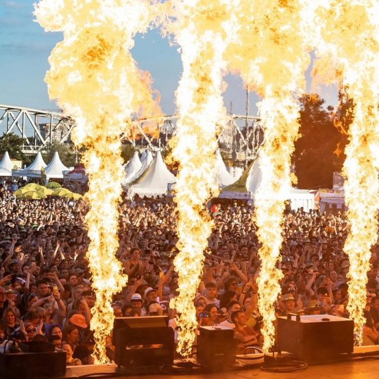 Eine große Menschenmenge versammelt sich um eine Bühne mit Lautsprechern und Pyrotechnik. Die Feuereffekte sind prominent und das Publikum ist aktiv beteiligt. Zelte und eine Brücke sind im Hintergrund zu sehen.
