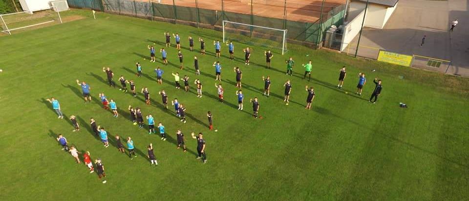 Eine Gruppe von Personen in Sportbekleidung führt Aufwärmübungen auf einem Rasenfeld durch, mit einem Fußballtor und einem Tennisplatz im Hintergrund.