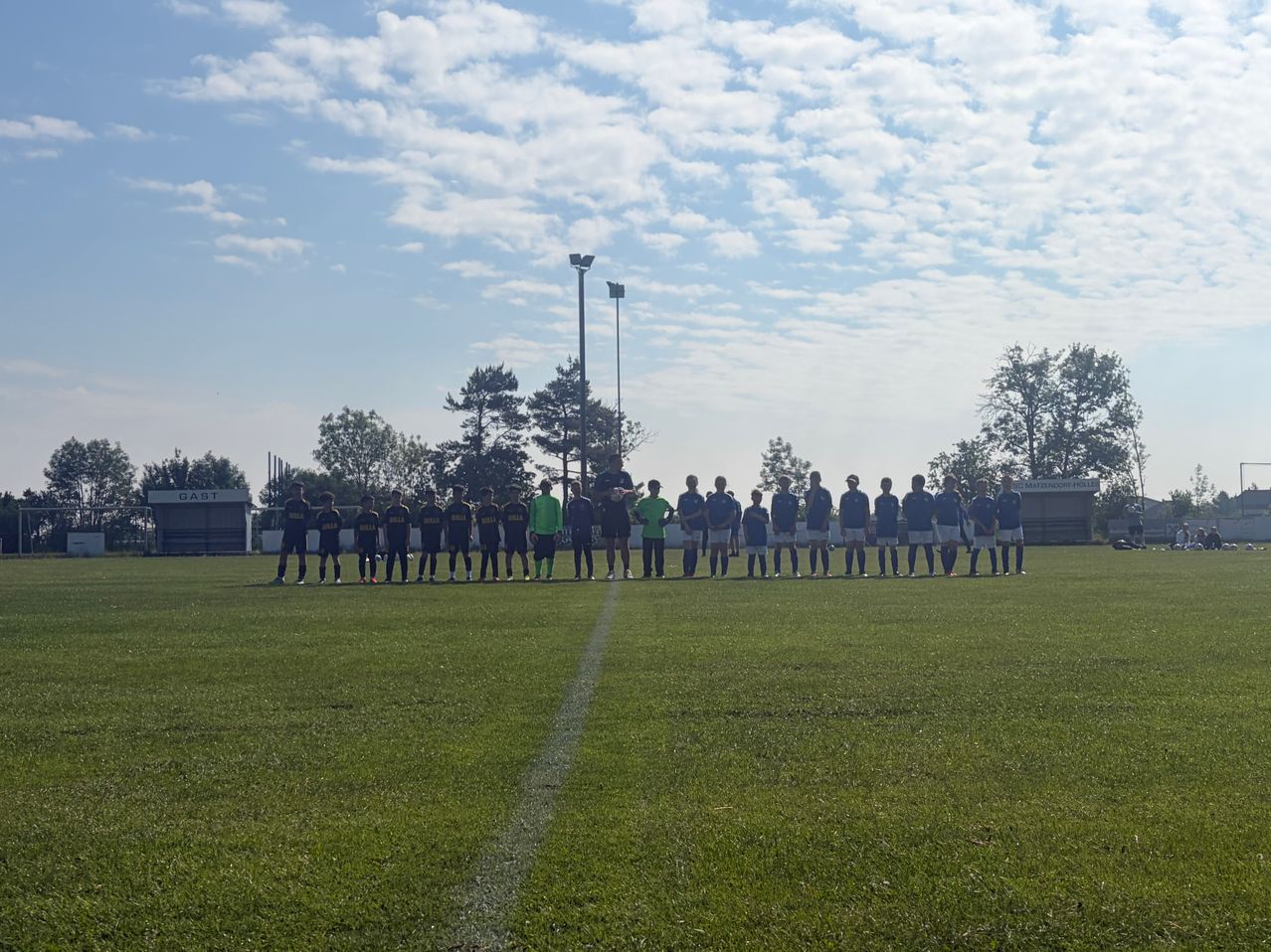 Eine Fußballmannschaft steht in einer Reihe auf einem Feld mit klarem blauem Himmel und Bäumen im Hintergrund.