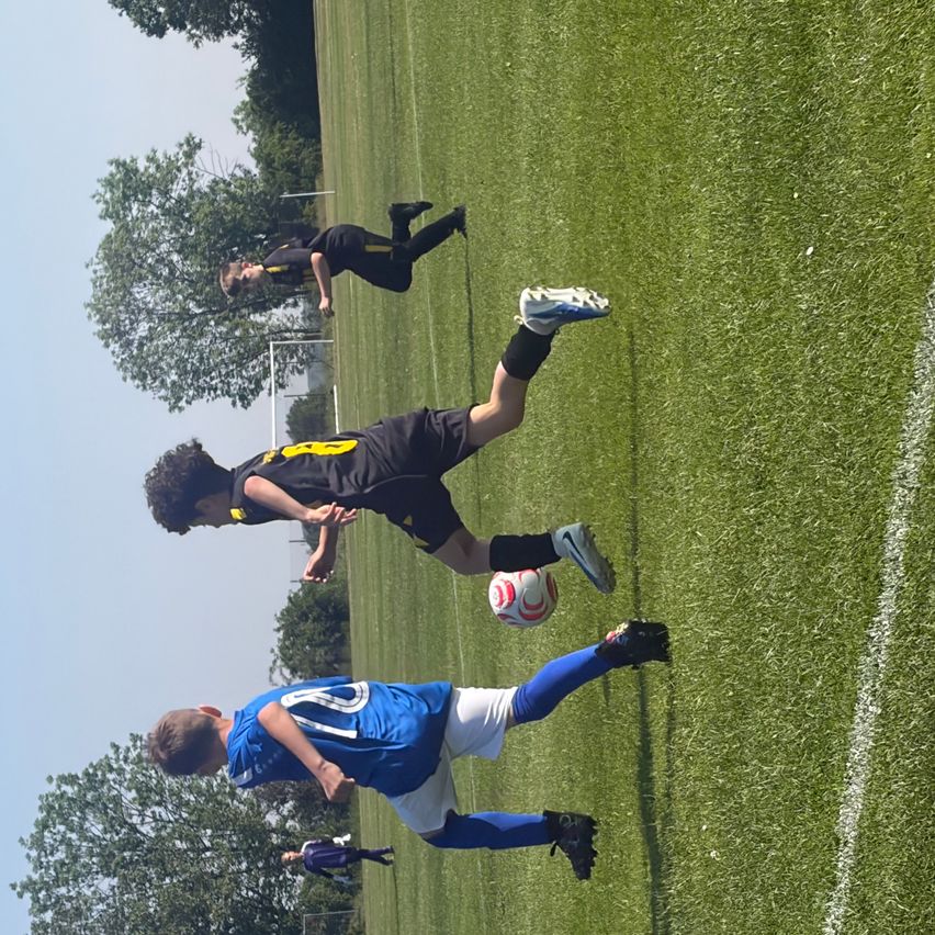Zwei Fußballspieler spielen auf einem Feld. Einer kickt den Ball, während der andere verteidigt. Das Feld hat weiße Linien und Bäume im Hintergrund.