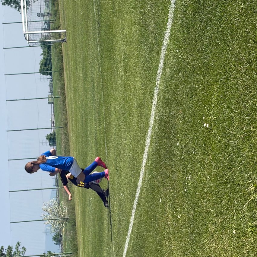 Eine Fußballspielerin in blauem Trikot versucht, den Ball zu treten, während sie von einer anderen Spielerin getackelt wird.