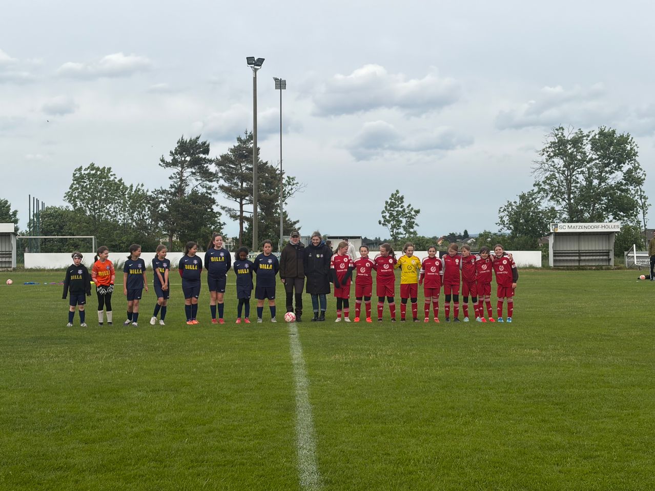 Eine Fußballmannschaft in roten und blauen Trikots steht auf einem Rasenfeld. Eine weiße Linie markiert das Feld, und Bäume sind im Hintergrund zu sehen.