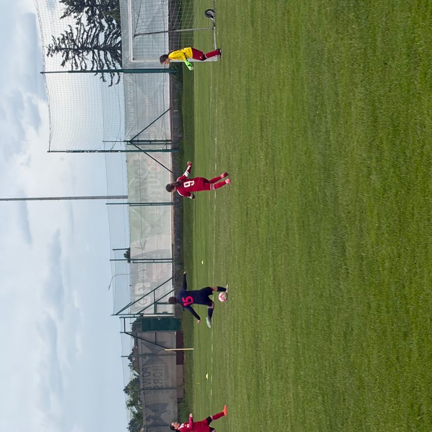 Luftaufnahme eines Fußballfeldes mit Spielern in roten, gelben und schwarzen Trikots. Spieler befinden sich auf dem Feld, einer in der Nähe des Tors.