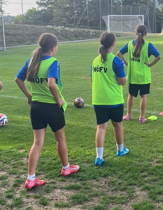 Drei Mädchen in grünen Westen stehen auf einem Fußballfeld und beobachten einen Ball. Sie tragen Turnschuhe und Shorts.