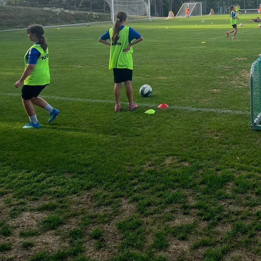 Zwei Mädchen in neongrünen Westen gehen auf einem Fußballfeld. Ein Fußball und Hütchen sind auf dem Feld.