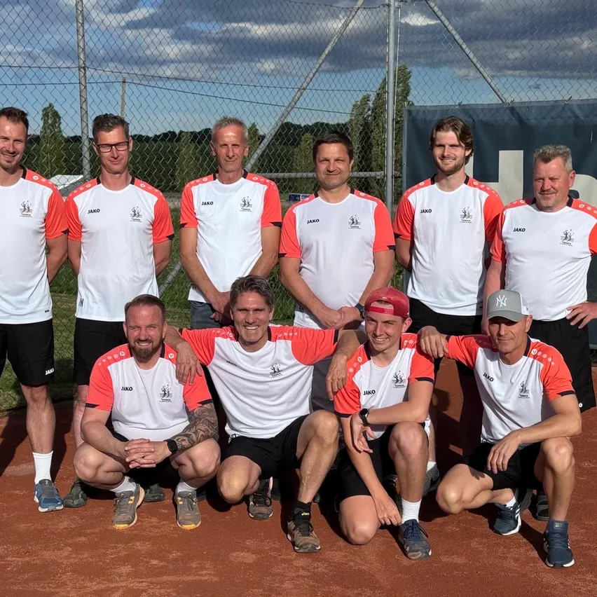 Eine Gruppe von Männern in gleichen Trikots posiert für ein Foto auf einem Tennisplatz, mit einem Zaun und Bäumen im Hintergrund.