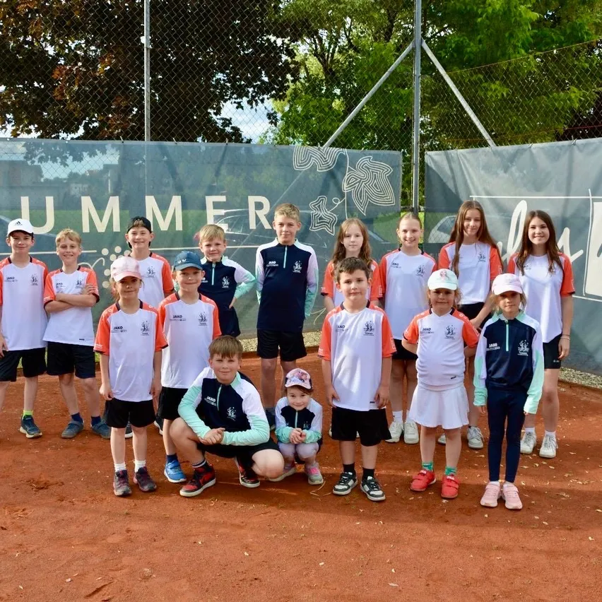 Eine Gruppe von Kindern in weißen und orangefarbenen Uniformen posiert für ein Foto auf einem Tennisplatz mit einem 'SUMMER'-Banner im Hintergrund.