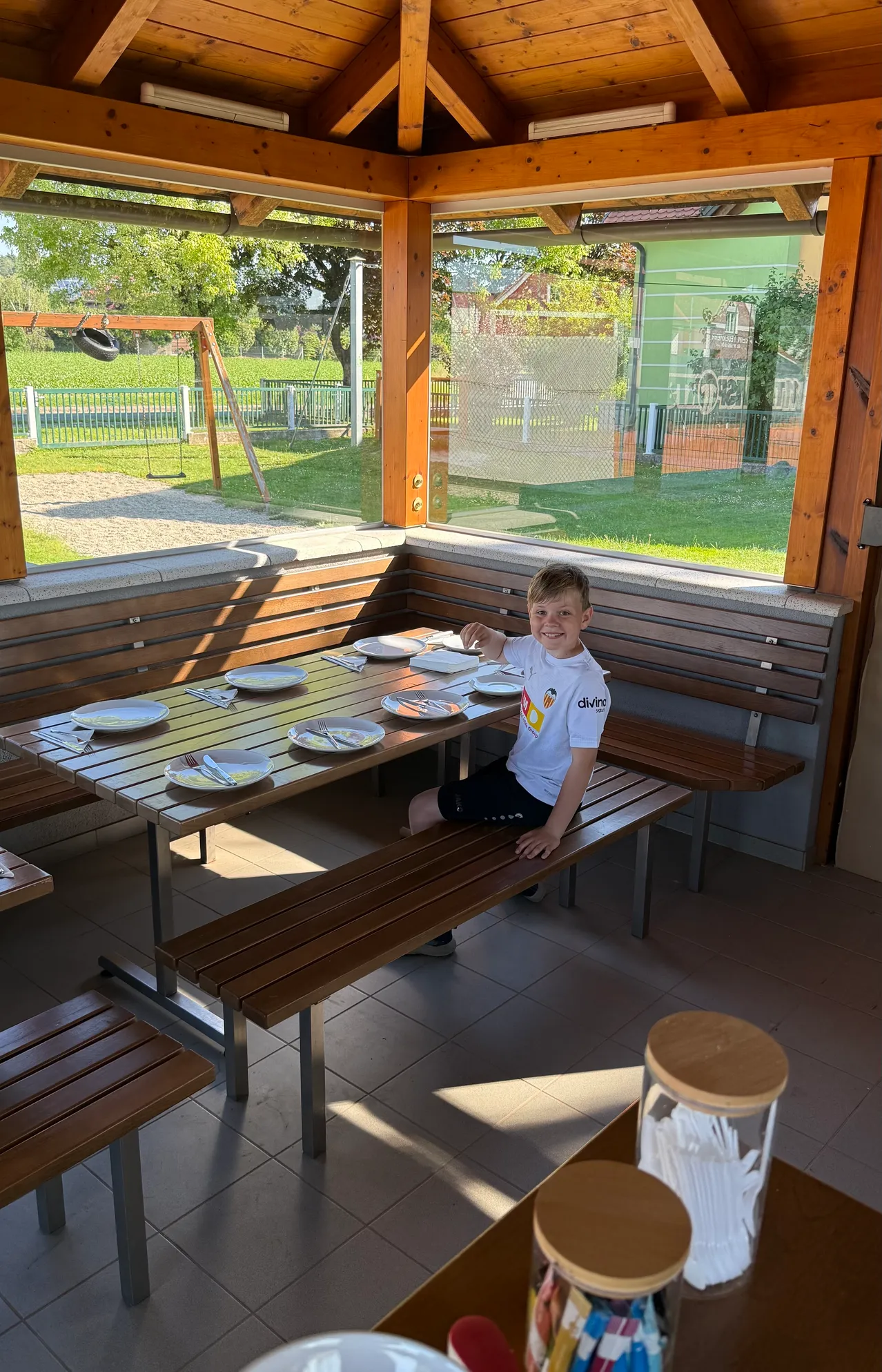Ein junger Junge in einem weißen T-Shirt sitzt an einem Tisch in einem Außenbereich. Auf dem Tisch liegen Teller, Besteck und eine Glasdose. Hinter ihm blickt ein Glasfenster auf einen Spielplatz mit Schaukel und einem Grasfeld.