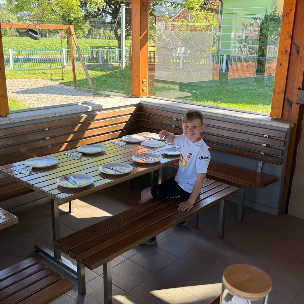 Ein junger Junge in einem weißen T-Shirt sitzt an einem Tisch in einem Außenbereich. Auf dem Tisch liegen Teller, Besteck und eine Glasdose. Hinter ihm blickt ein Glasfenster auf einen Spielplatz mit Schaukel und einem Grasfeld.