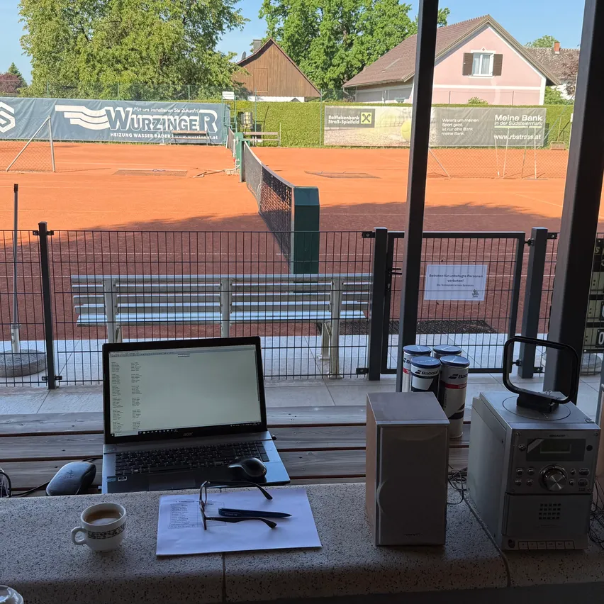 Ein Laptop, Kaffeetasse, Stift und eine Brille auf einem Tresen vor einem Tennisplatz. Ein geöffneter Laptop zeigt einen Bildschirm an.