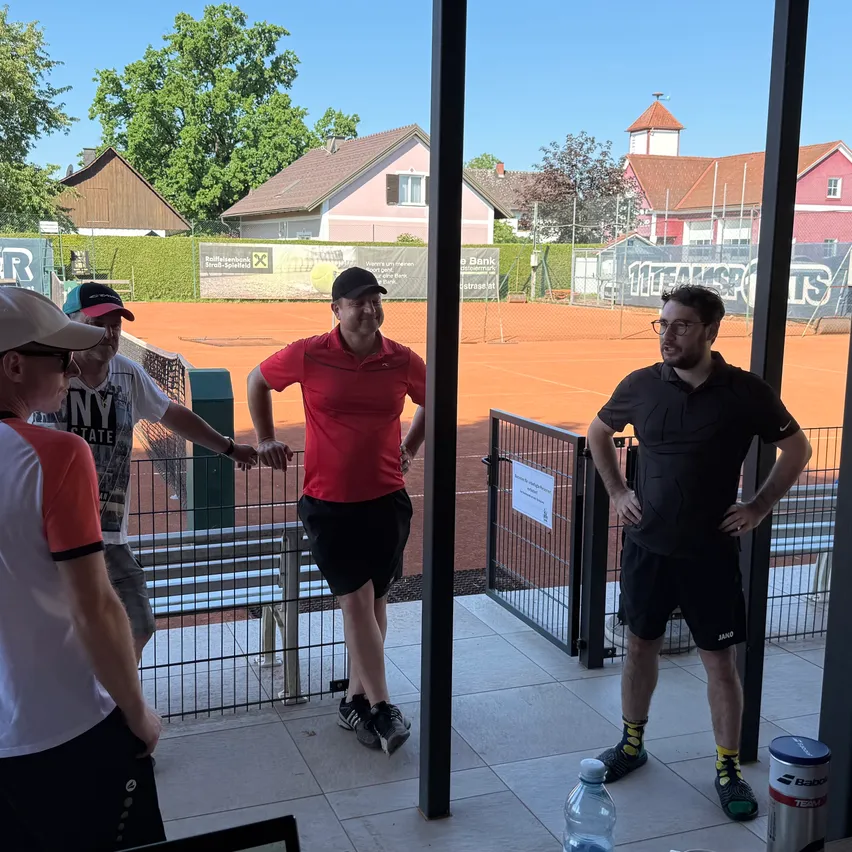 Vier Männer stehen am Eingang eines Tennisplatzes. Sie tragen Hüte und Turnschuhe. Ein Mann in der Mitte trägt ein rotes Shirt und schwarze Shorts. Eine Flasche und ein Wasserspender stehen auf dem Boden. Dahinter befindet sich ein Tennisplatz mit einem Zaun.