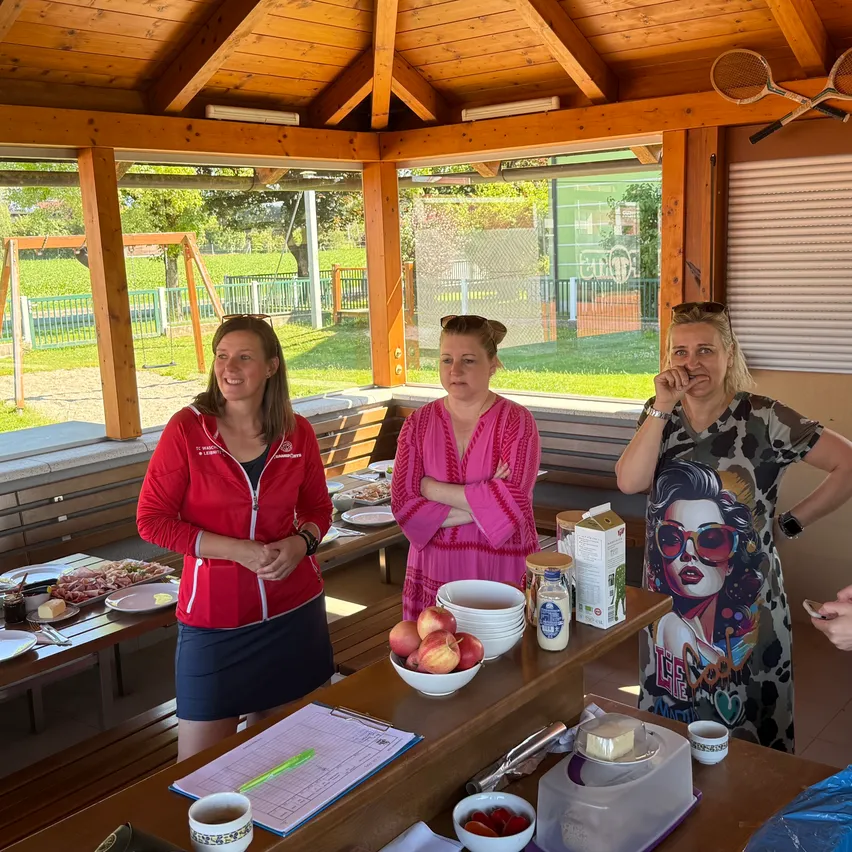 Drei Frauen stehen um einen Tisch in einem hölzernen Pavillon. Sie lächeln und scheinen Essen zuzubereiten. Auf dem Tisch stehen Schüsseln mit Obst, Teller und eine Milchpackung. Der Pavillon befindet sich in der Nähe eines Spielplatzes.