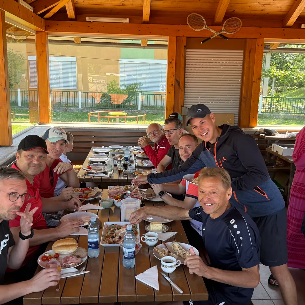 Eine Gruppe von Männern sitzt um einen Tisch herum und lächelt für ein Foto. Der Tisch ist mit Tellern mit Essen, Tassen und Flaschen gedeckt. Hinter ihnen gibt es ein großes Fenster und eine Aussicht auf einen Rasenbereich mit einem Zaun.