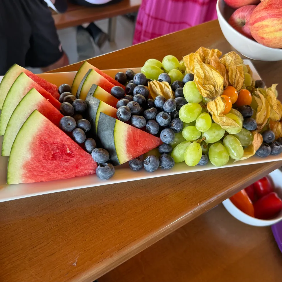 Ein Obstplatten auf einem Holztisch mit Wassermelone, Blaubeeren, Trauben und getrockneten Blumen. Im Hintergrund befinden sich Schalen mit Äpfeln und Tomaten.