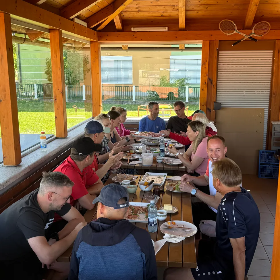 Eine Gruppe von Menschen sitzt um einen Tisch in einem Pavillon und genießt eine Mahlzeit. Sie sind lässig gekleidet, einige tragen Mützen. Der Tisch ist mit Tellern mit Essen und Getränken gedeckt. Ein Tennisschläger ist an der Wand hinter ihnen montiert.