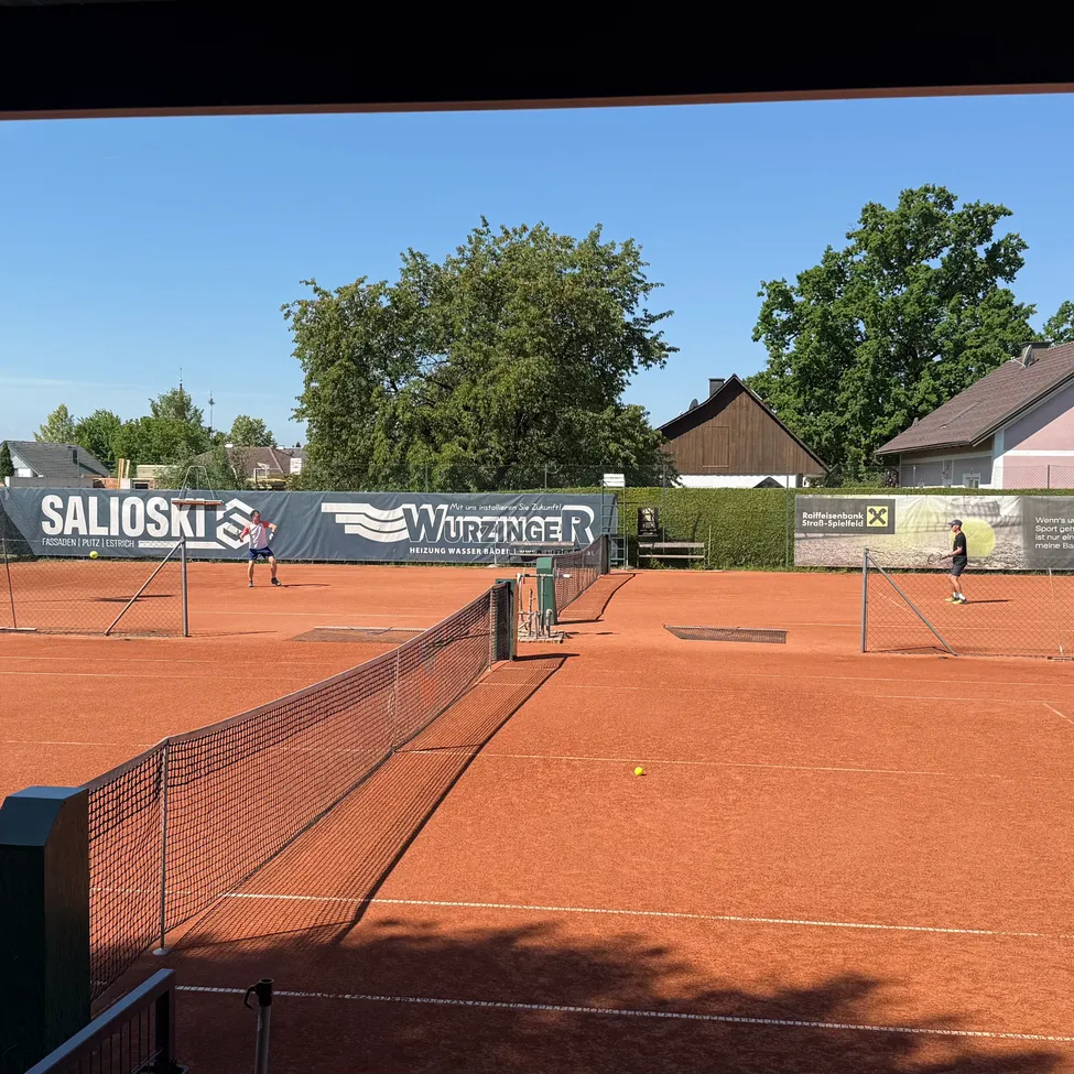 Zwei Spieler spielen Tennis auf einem Sandplatz. Einer ist in der Nähe des Netzes und der andere hinten. Der Platz ist von einem Zaun umgeben. Im Hintergrund sind Bäume und Häuser zu sehen.