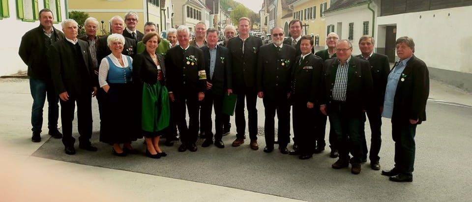 Eine Gruppe formell gekleideter Personen, einige in traditioneller Kleidung, steht vor Gebäuden in einem Dorf.