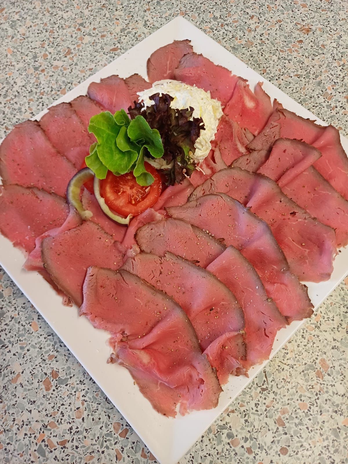 Ein weißer Teller mit geschnittenem Roastbeef, garniert mit einer Tomate, Salat und einem Klecks Frischkäse.