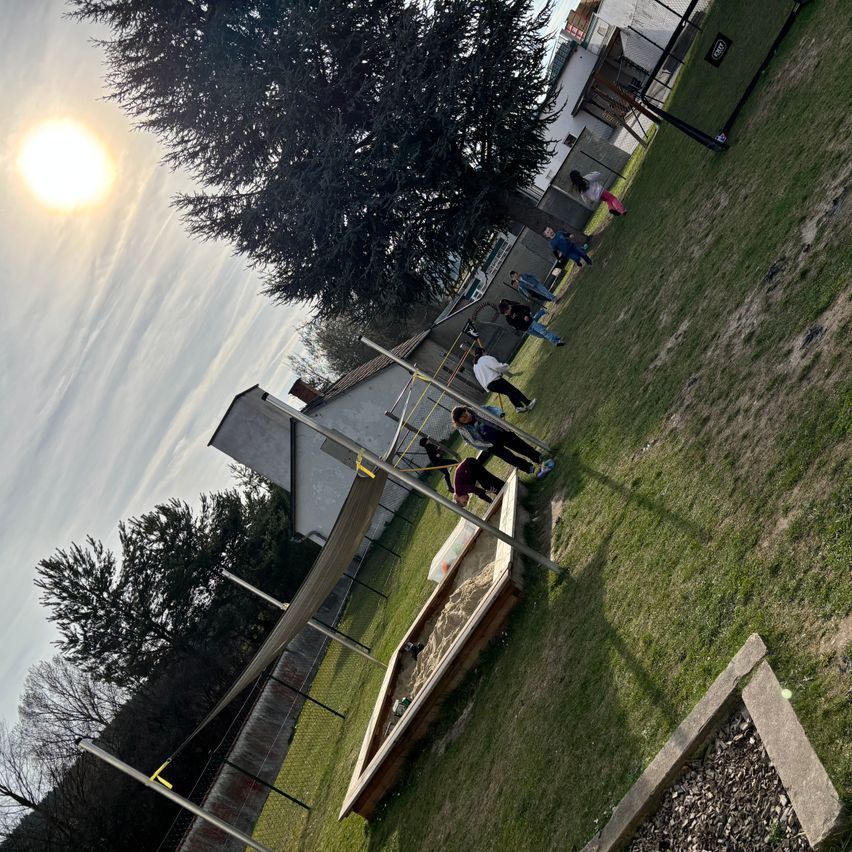 Ein sonniger Tag mit Kindern, die auf einem Spielplatz mit Sandkasten spielen, umgeben von Bäumen und Gebäuden.