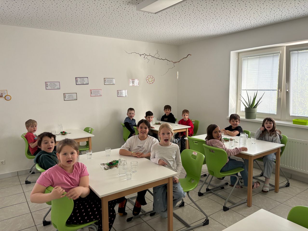 Ein Klassenzimmer mit Kindern, die an Tischen sitzen. Ein Mädchen in Pink trägt einen gepunkteten Rock. Ein Ast hängt von der Decke, und eine Topfpflanze ist am Fenster.