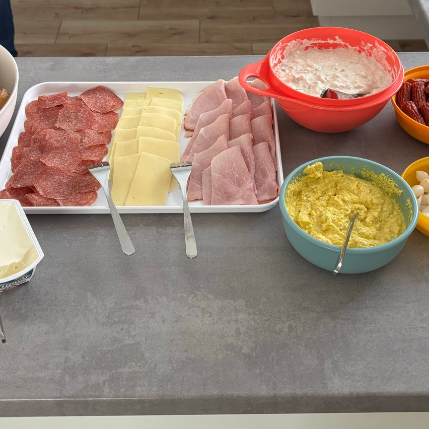Ein Küchentisch zeigt verschiedene Lebensmittel, darunter geschnittene Salami, Schinken, Käse und zwei Schüsseln mit Dips. Zwei Gabeln liegen neben den Lebensmitteln auf einem Tablett. Auf der rechten Seite des Tisches befinden sich eine Schüssel mit roter Soße und eine weitere mit gelber Soße.