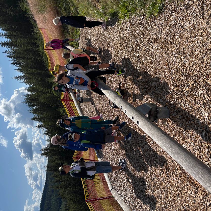 Eine Gruppe von Kindern und Erwachsenen geht auf einem Holzweg mit einem gelb-roten Zaun, umgeben von Bäumen und einem blauen Himmel mit Wolken.