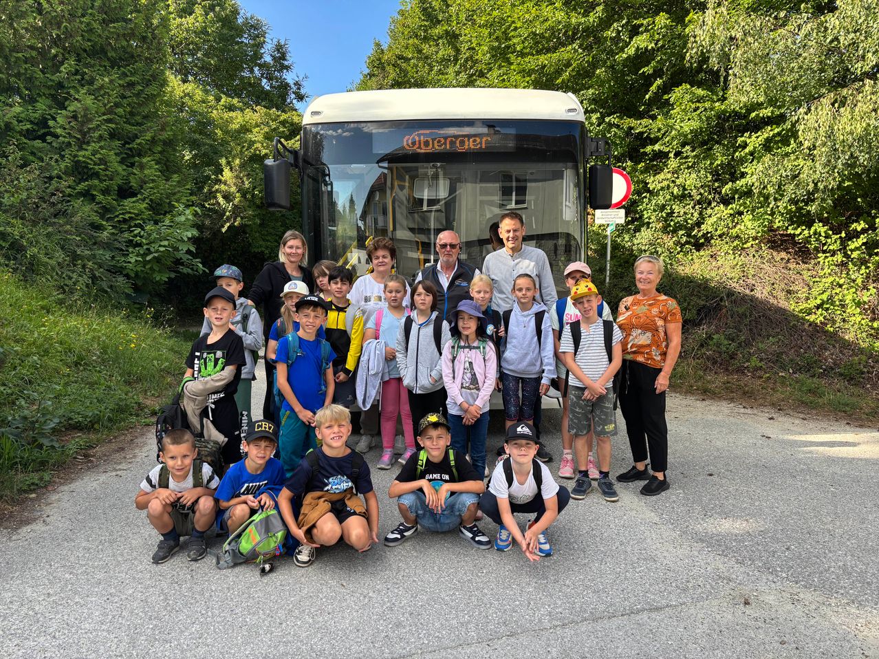 Eine Gruppe von Kindern und Erwachsenen posiert für ein Foto vor einem Bus mit dem Wort 'Berger' darauf. Sie stehen und sitzen auf dem Boden, einige tragen Hüte und haben Rucksäcke dabei.