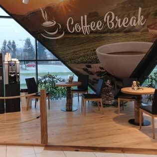 Ein Café mit einem Wandbild, das Kaffee Pause sagt. Es gibt Tische und Stühle, eine Kaffeemaschine und eine Tasse Kaffee auf einem großen Poster.