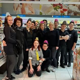 Eine Gruppe von Menschen in schwarzer Kleidung und festlichen Hüten posiert für ein Foto in einem Lebensmittelgeschäft mit verschiedenen Produkten im Hintergrund.