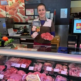 Eine Frau in einem braunen Shirt steht an einer Fleischtheke, hält ein Buch und ein Stück Fleisch. Hinter ihr ist eine Theke mit Gemüse und einem Monitor. Die Theke zeigt verschiedene Fleischsorten an.