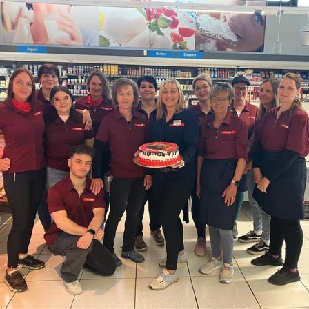 Eine Gruppe von Mitarbeitern in roten Uniformen posiert mit einem Kuchen in einem Supermarkt. Eine Frau hält einen Kuchen, und ein Mann kniet davor. Hinter ihnen sind Regale mit Lebensmitteln zu sehen.