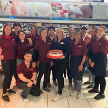 Eine Gruppe von Mitarbeitern in roten Uniformen posiert mit einem Kuchen in einem Supermarkt. Eine Frau hält einen Kuchen, und ein Mann kniet davor. Hinter ihnen sind Regale mit Lebensmitteln zu sehen.