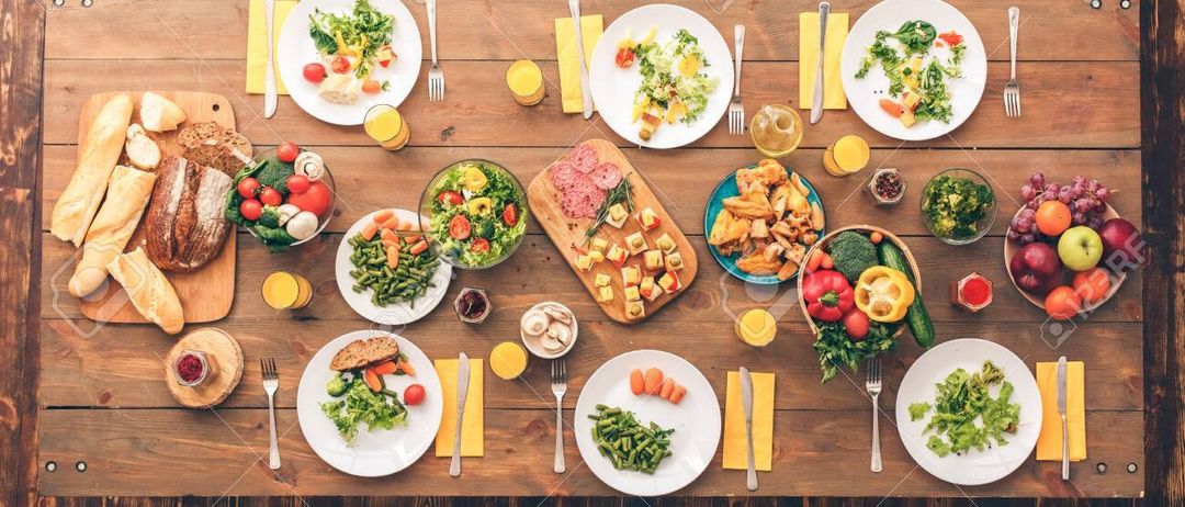 Ein Holztisch mit mehreren Tellern verschiedener Gerichte, darunter Salate, Gemüse und Brot, für eine Mahlzeit vorbereitet. Die Teller sind in einer Reihe angeordnet, jeder mit einer Gabel und einem Messer. Zusätzliche Gegenstände wie Orangensaft und Gewürze sind auf dem Tisch platziert.