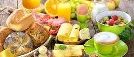 Ein Holztisch ist mit verschiedenen Frühstücksspeisen gedeckt, darunter Brot, Käse, Orangensaft, eine Obstschüssel und eine Tasse Kaffee.