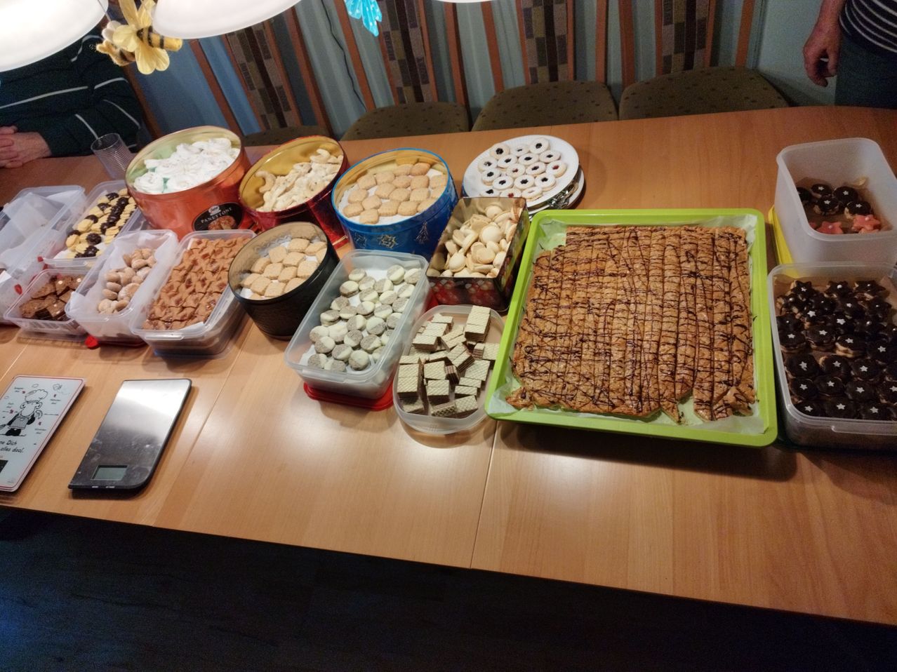A wooden table displays an assortment of cookies in various containers, including a tray with chocolate drizzle. A scale is placed on the left side.