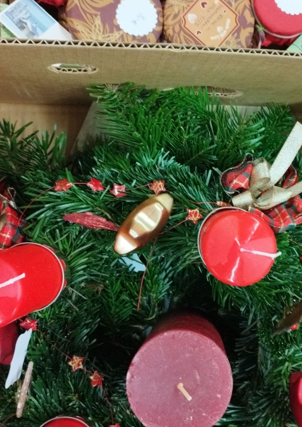 Eine Nahaufnahme eines Weihnachtskranzes mit roten Kerzen, einem goldenen Ornament und roten Sternen auf einem grünen Zweig. Der Kranz befindet sich in einer Pappschachtel.